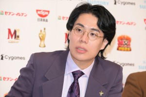 紫色のスーツと紫のネクタイを着用し丸い眼鏡をかけた黒髪の若い男性が会見やイベントに出席している様子 企業ロゴが背景に並んでいる
